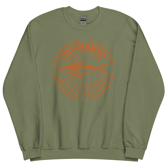 Sudadera verde militar, con un logotipo naranja en el centro. El logotipo representa a un chicharro dibujado de lado con la forma abierta. El chicharro está rodeado por un círculo, en el que aparecen las palabras CHICHARRO, TATTO GALLERY, CANARY ISLANDS y SANTA CRUZ DE TENERIFE.